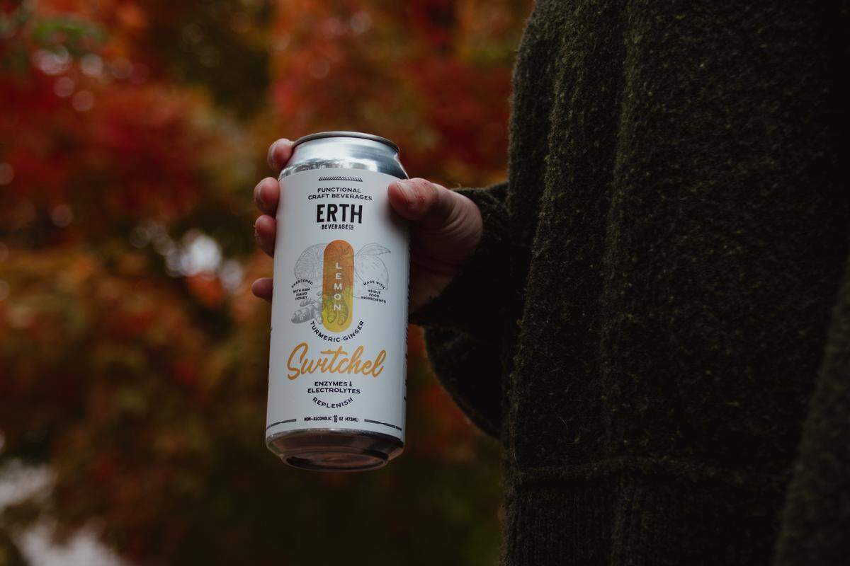 A hand holding a can labeled as a Lemon Turmeric switchel in front of a backdrop of autumn leaves.