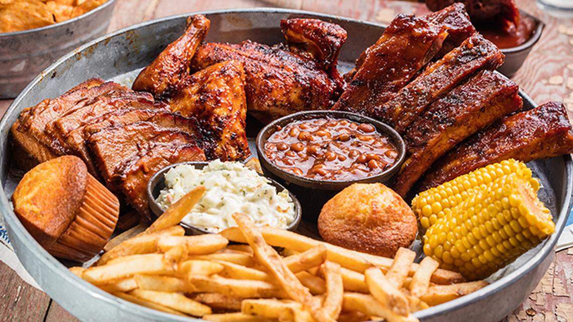 A mouth-watering barbecue feast served on a round metal platter. The platter is piled high with glistening ribs, smoked chicken, crispy french fries, corn on the cob, golden cornbread muffins, baked beans, and creamy coleslaw, creating an abundant and delicious-looking meal.