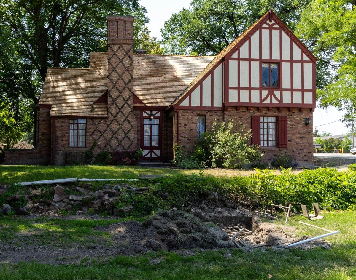 The historic Orville Jackson House, with its characteristic brick and stucco exterior, as seen from Eagle Road in downtown Eagle in June 2025. From the rock wall, the area around the home is protected by a historical easement. The home is preserved by a deed restriction preventing demolition in perpetuity. Exposed piping in the foreground shows the extent of some repairs needed.