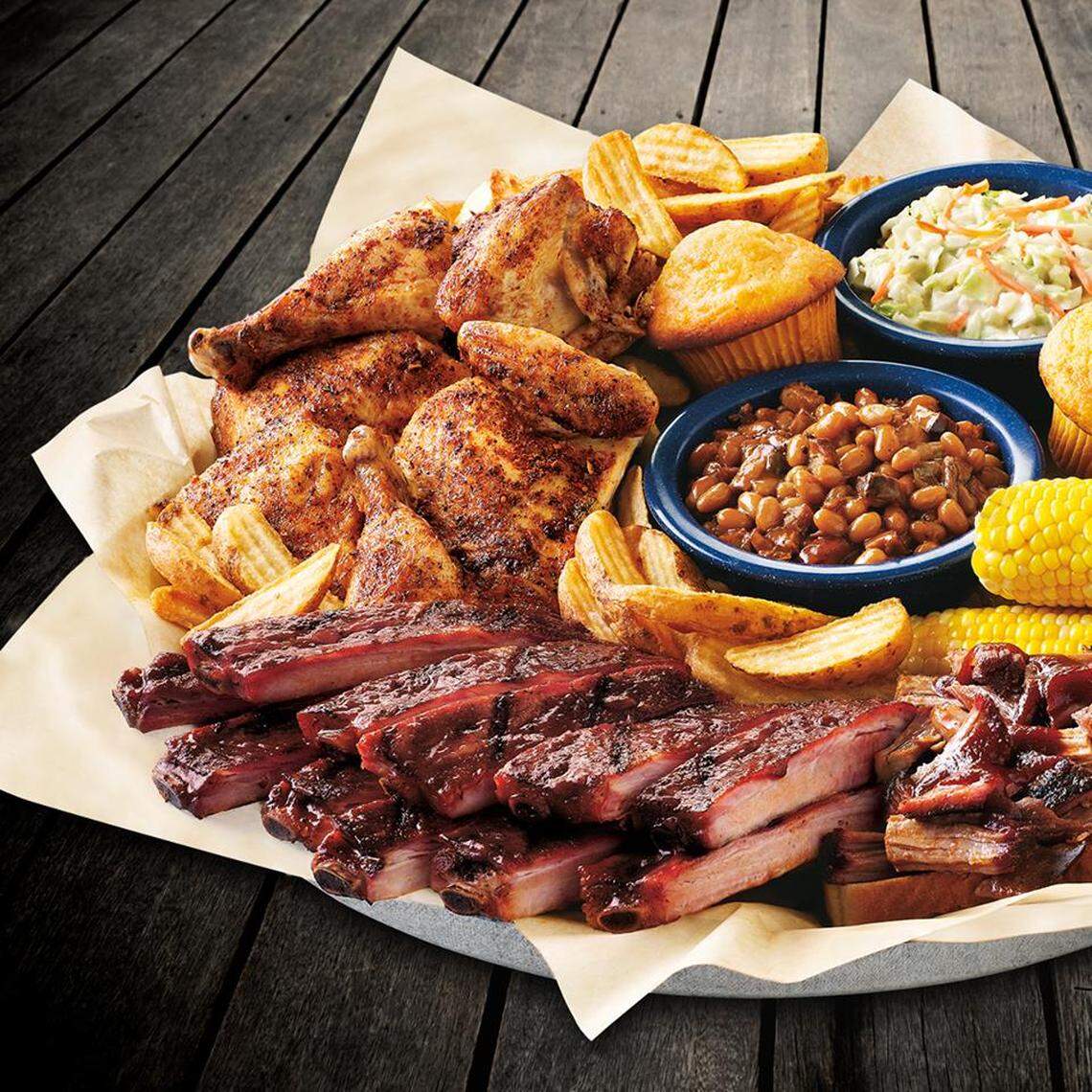 A bird’s-eye view of an abundant barbecue platter resting on a dark wood-plank surface. The platter is loaded with a seasoned cut-up chicken, a row of glossy barbecue ribs, potato wedges, cornbread muffins, corn on the cob, and blue bowls of baked beans and coleslaw.