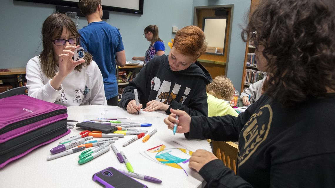 While visiting Teenspace at the Meridian Library after school, Amber Bell, 12, left, holds up her drawing on plastic during a Shrinky Dink craft project with Chandler Ston, 13, and Takya Sanabria, 12, right. Program specialist Pamela Hay, back, helps the teens with heating the Shrinky Dinks, which are polystyrene shapes that shrink and harden when heated.