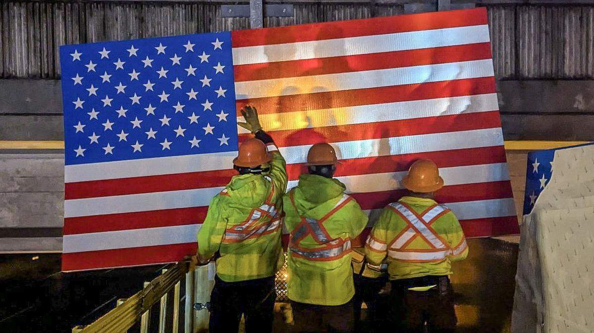 Crews with the Idaho Transportation Department hang an American flag sign on the Cloverdale Road overpass above I-84 in Boise in April 2026. The sign memorializes three Idaho airmen who died in a fiery crash in 2018 on the Interstate below.
