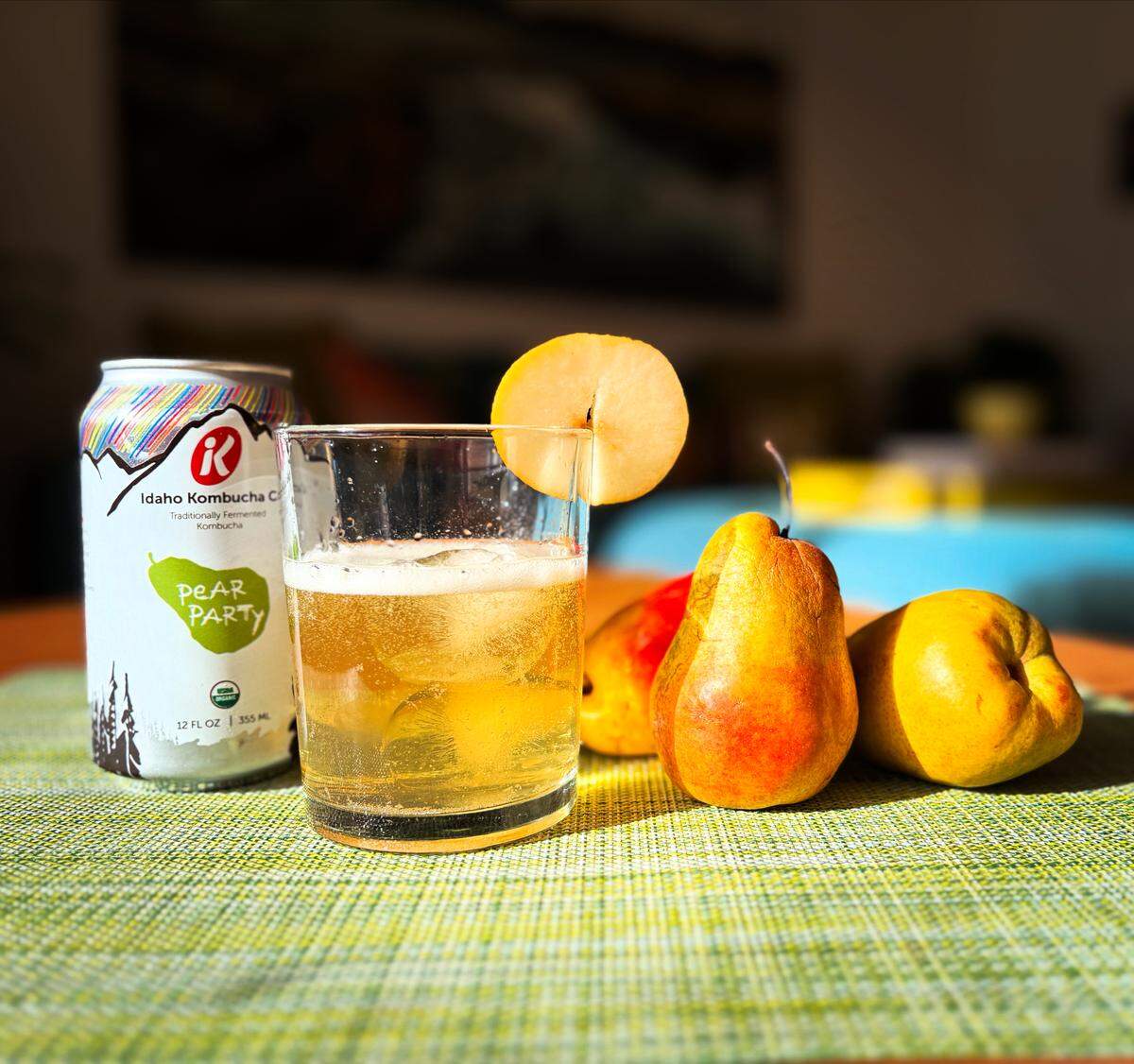 A can of Pear Party kombucha from the Idaho Kombucha Co next to a glass of kombucha with a pear slice on the rim and a few pears surrounding the glass.