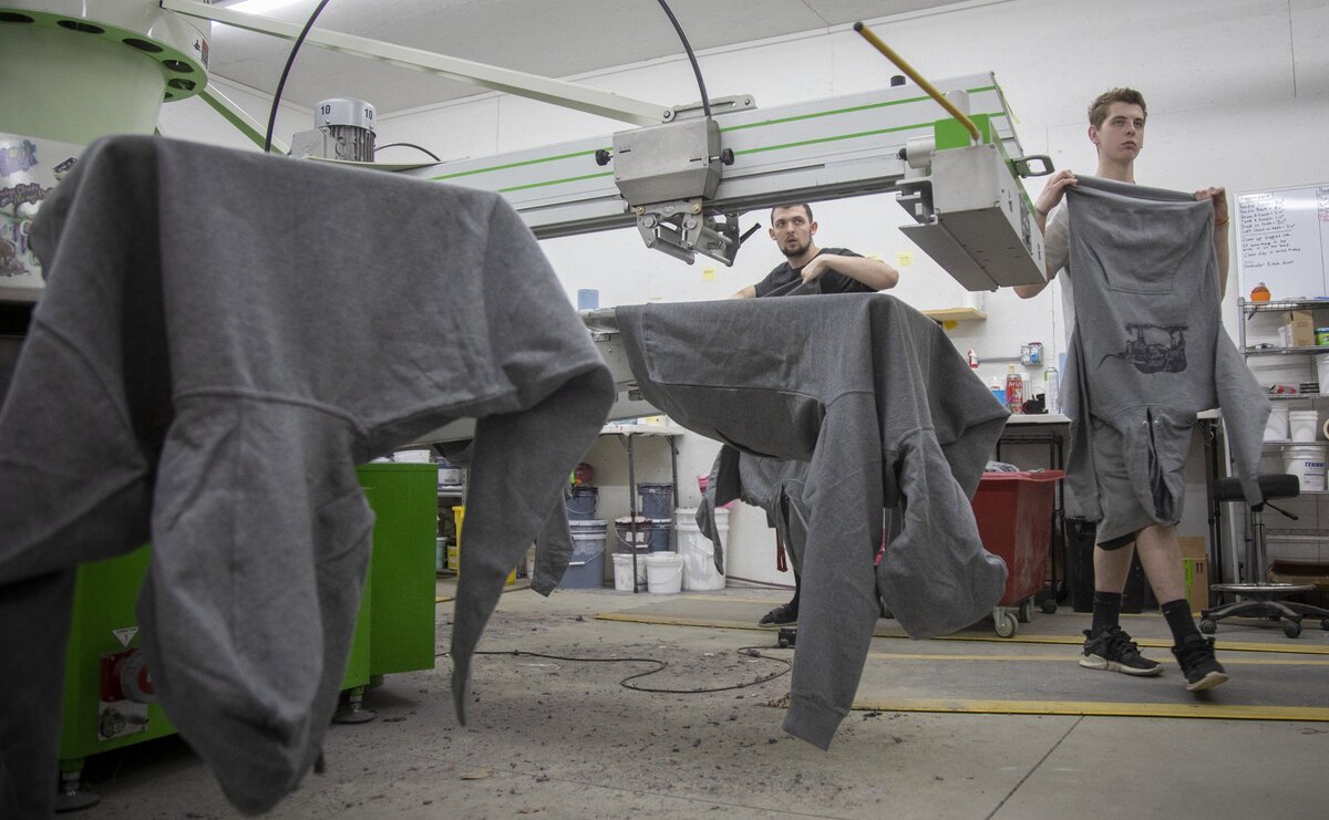 Jessi Roberts’ family is part of Cheekys, including her son, Hunter Crites, center, who smooths t-shirts on to the 12-palette screen printing press, and his best friend Tristan Echardt, who pulls them off and puts them into the drier.