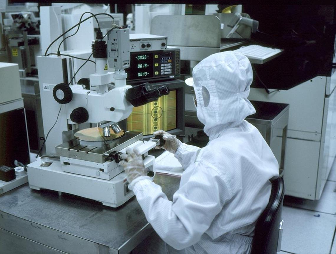 A Micron worker in a “bunny suit” inspects a silicon wafer upon which memory chips are etched in a grid.