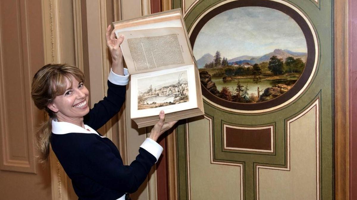 Amy Burton holds the Pacific Railroad Survey volume, where she found the Old Mission lithograph. The Cataldo landscape medallion is on the wall behind her.